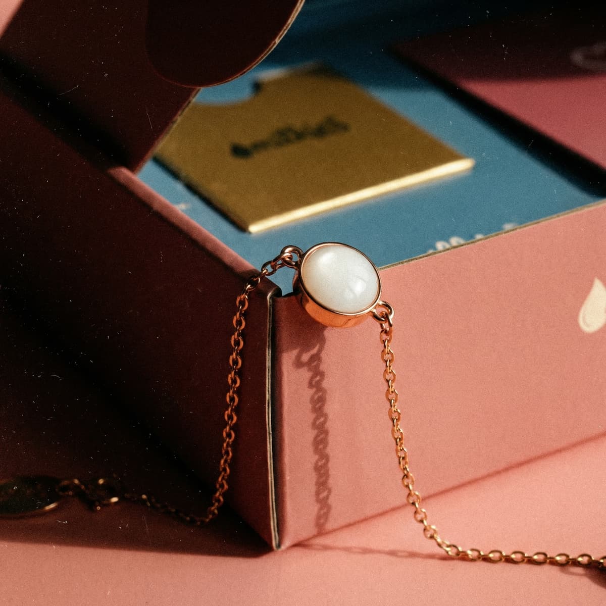 Pozłacana bransoletka z mlekiem matki DIY z białym kaboszonem w oprawie, ułożona na różowym pudełku zestawu do tworzenia pamiątkowej biżuterii w domu.