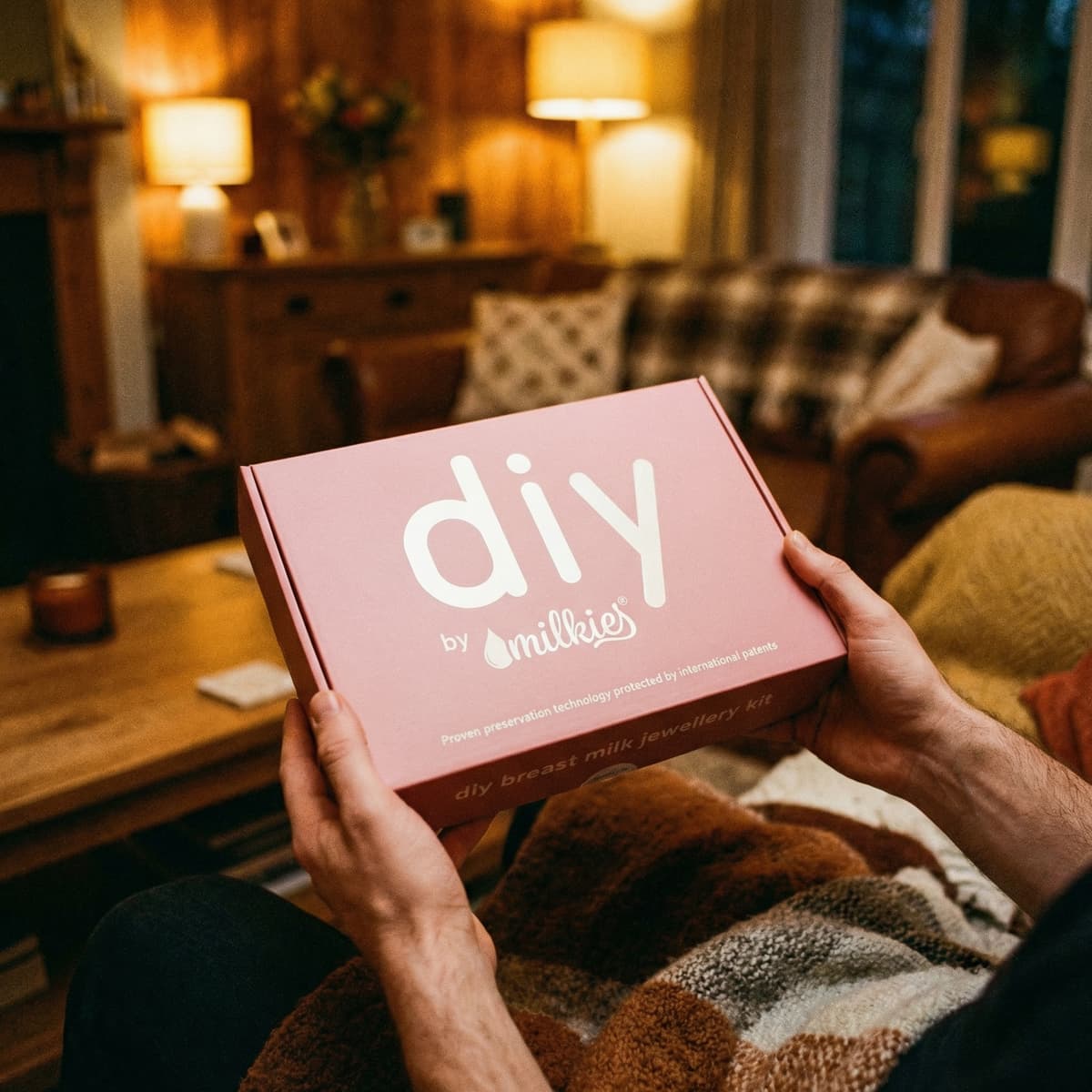 Domowa atmosfera: dłonie trzymają różowe pudełko DIY by MILKIES, zestaw do wykonania biżuterii z mleka mamy jako prezent dla żony za urodzenie dziecka.