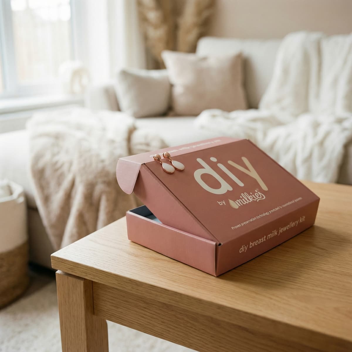 Elegancki zestaw DIY by MILKIES do tworzenia biżuterii z mleka mamy w różowym pudełku na drewnianym stoliku w jasnym salonie, idealny jako prezent na koniec karmienia.