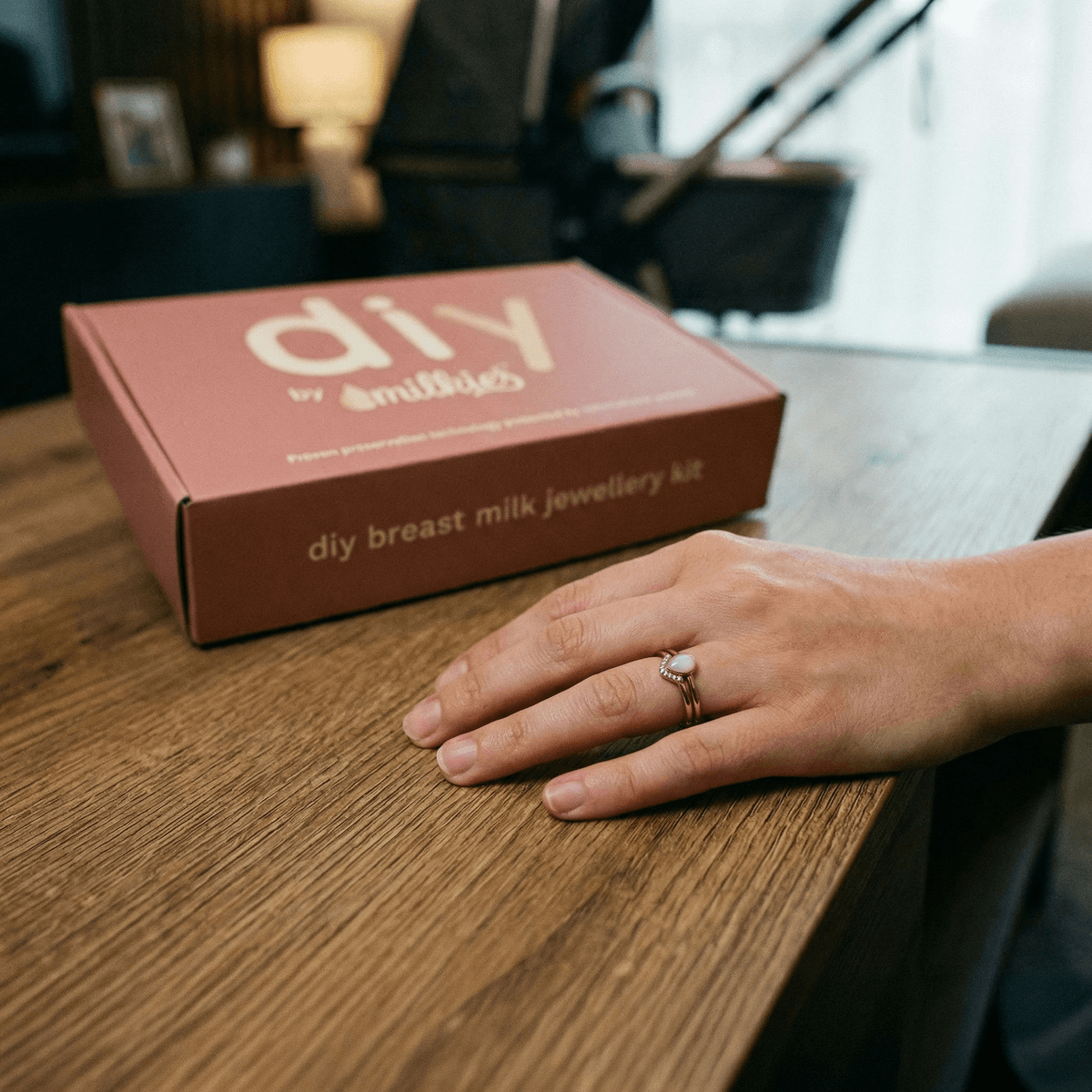 Dłoń kobiety z pierścionkiem z mleka mamy obok pudełka DIY by MILKIES na drewnianym stole, personalizowana biżuteria DIY jako wyjątkowy prezent na narodziny.