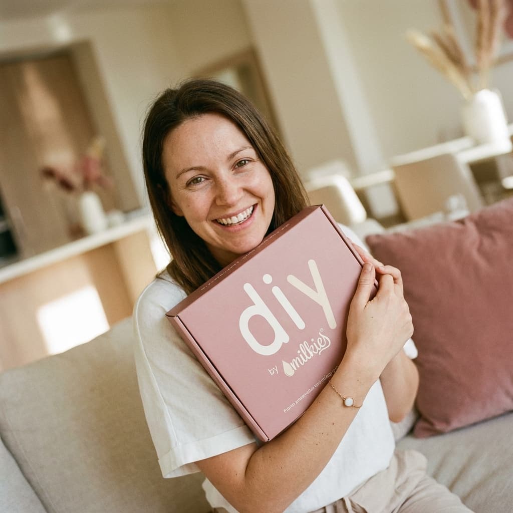 Uśmiechnięta mama w domu trzyma różowe pudełko DIY by MILKIES, pokazując dlaczego zestaw bransoletka z mleka matki to wygodny i dyskretny sposób na samodzielne stworzenie wyjątkowej pamiątki.