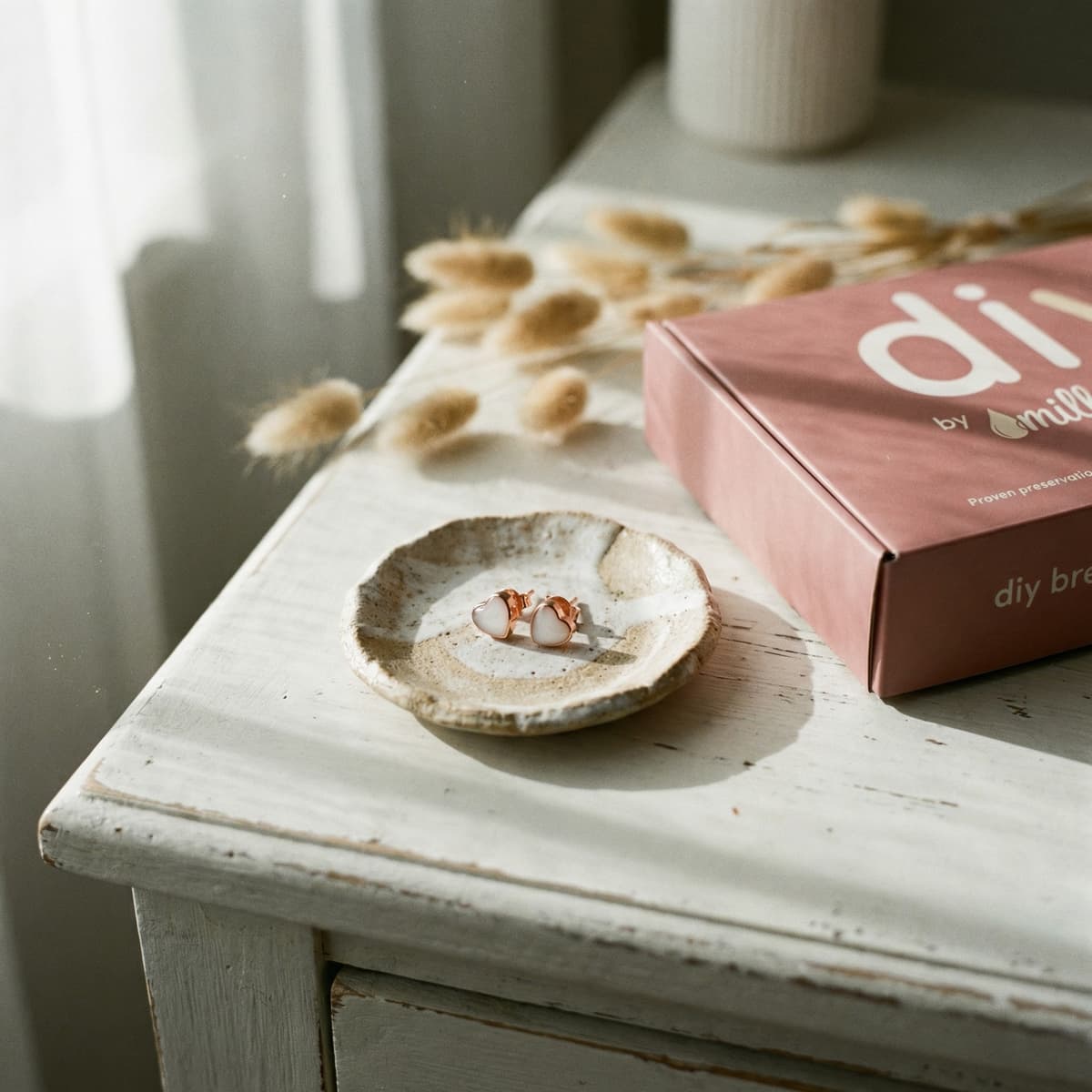 Na białym stoliku stoi różowy zestaw DIY by MILKIES, czyli zestaw do robienia kolczyków z mleka, a obok na ceramicznej miseczce leżą gotowe kolczyki w złotej oprawie z mlecznobiałym kamieniem.