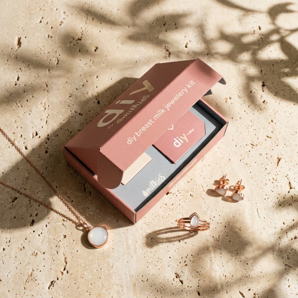 Różowe opakowanie DIY by MILKIES i przykładowa biżuteria (naszyjnik, pierścionek i kolczyki) z białym kamieniem prezentujące zestaw do tworzenia biżuterii z mleka matki na jasnym kamiennym tle.