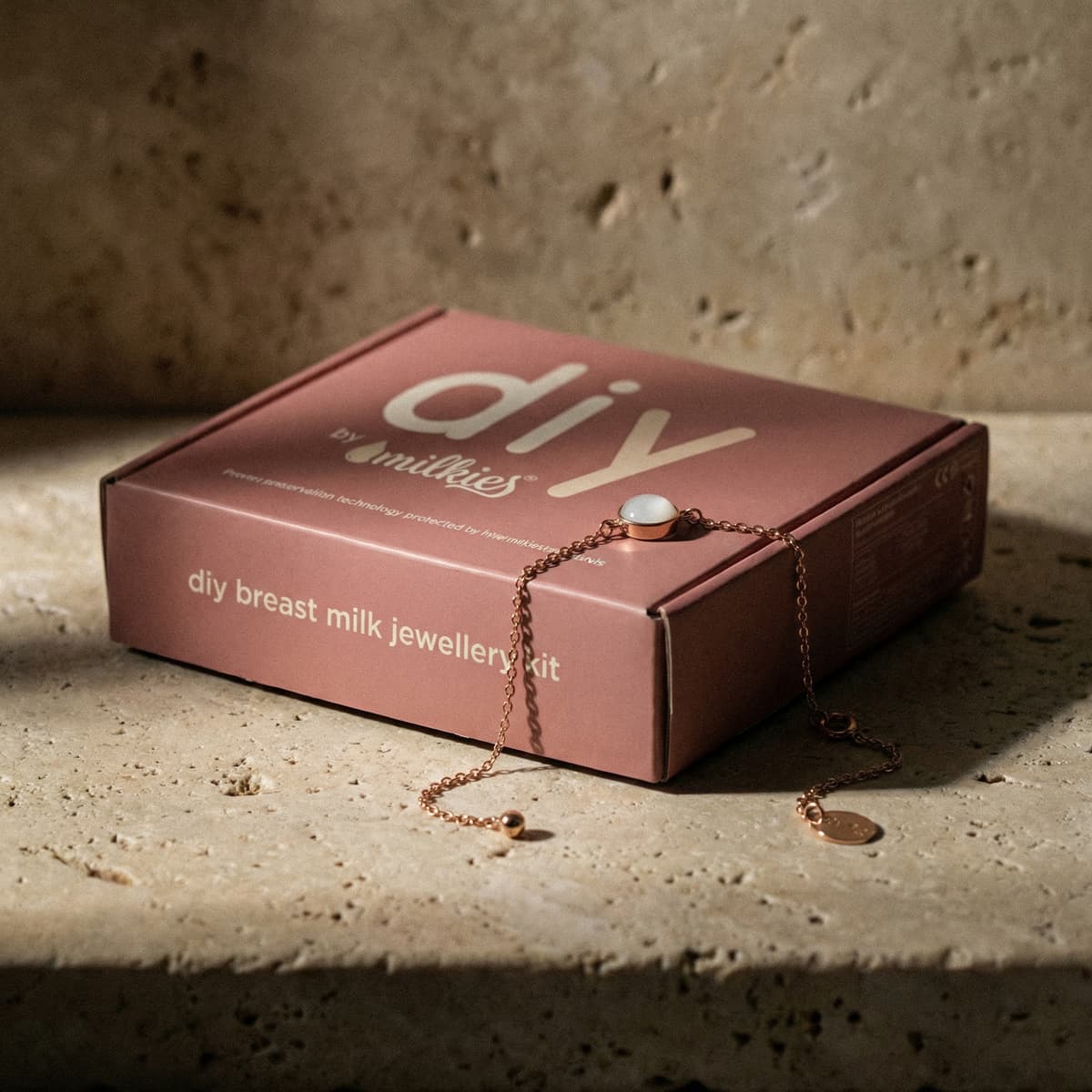 Różowe opakowanie DIY by MILKIES z napisem „diy breast milk jewellery kit” oraz delikatna bransoletka na łańcuszku z białym kamieniem, czyli zestaw do zrobienia bransoletki z mleka do samodzielnego wykonania pamiątkowej biżuterii