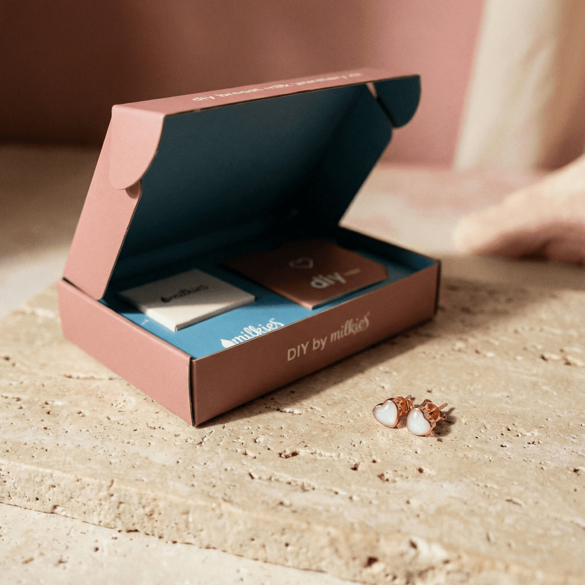 Otwarte pudełko DIY by MILKIES na kamiennym blacie z elementami do wykonania pamiątki oraz zestaw kolczyki z mlekiem matki w kształcie serduszek w różowo‑złotej oprawie.